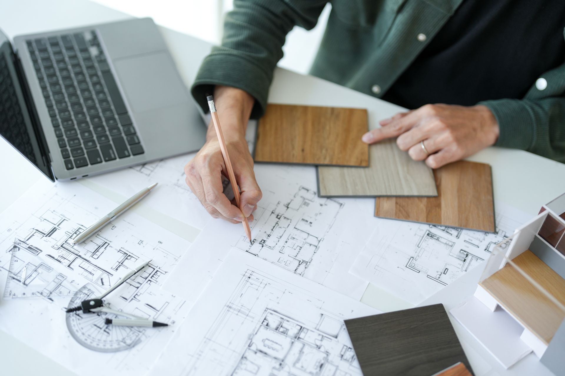 Person sketching architectural plans, selecting wood samples, laptop and tools on desk.