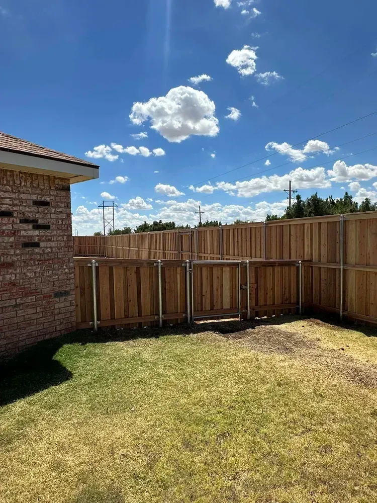 Backyard with a wooden fence, brick house wall, and sunny blue sky with clouds