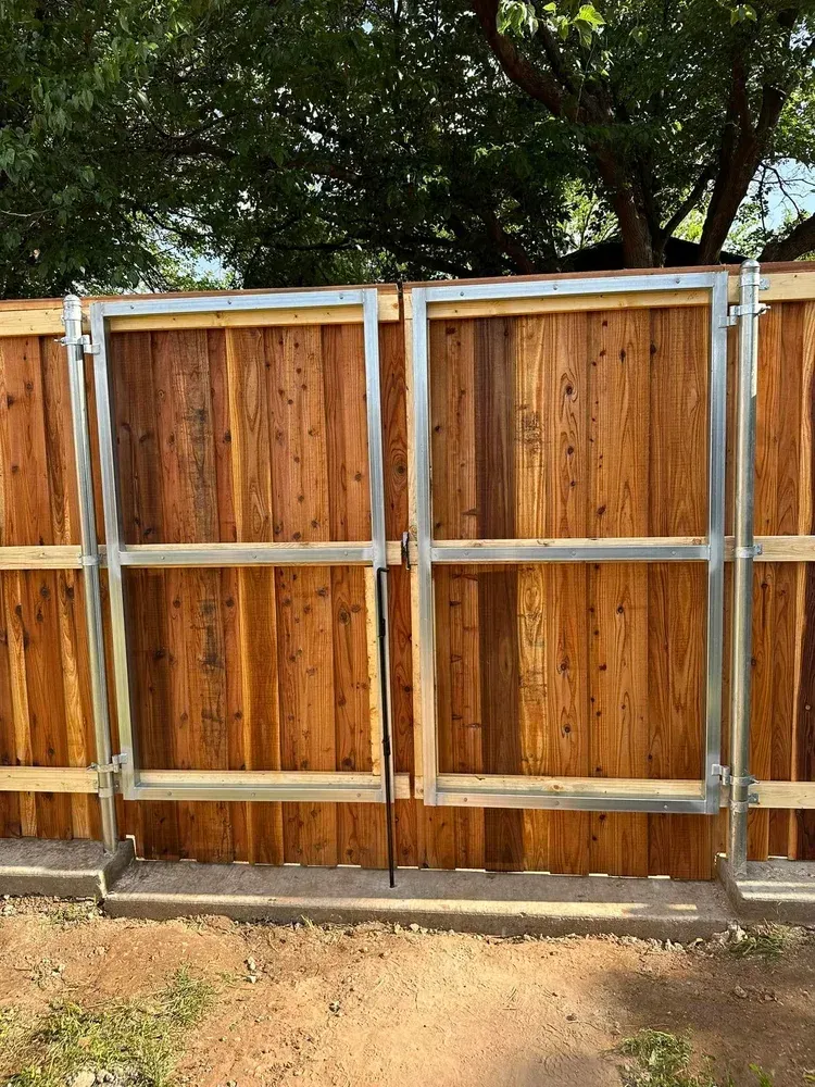 Wooden privacy fence with gray metal support rails in a backyard, with trees overhead and dirt ground below.