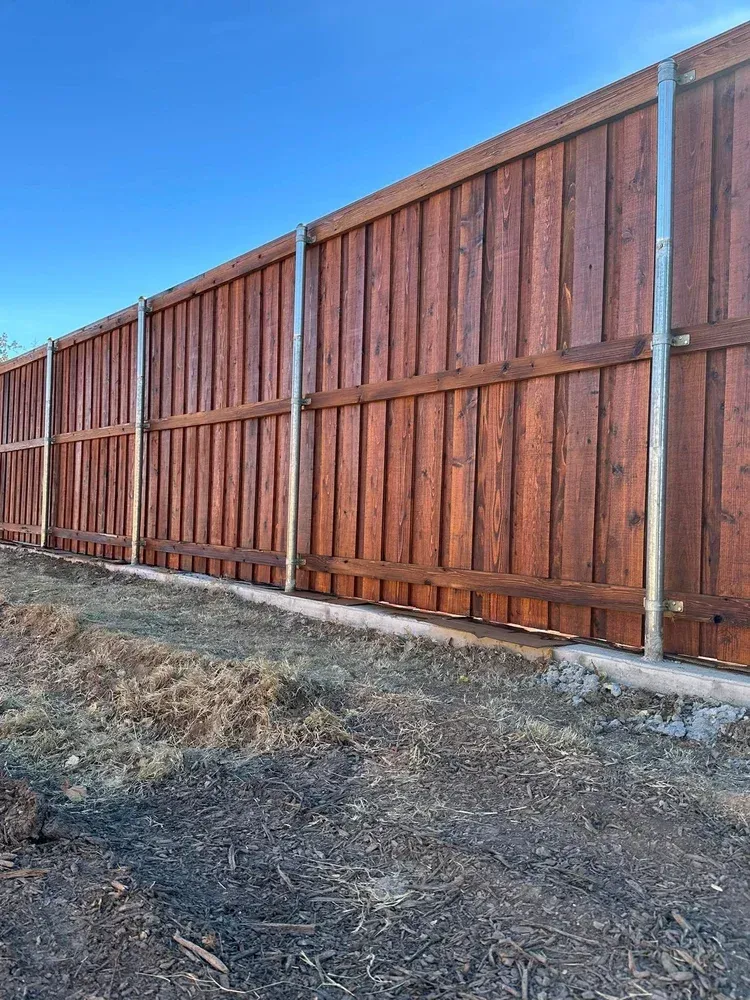 Tall brown wooden privacy fence along a dirt roadside under a clear blue sky.