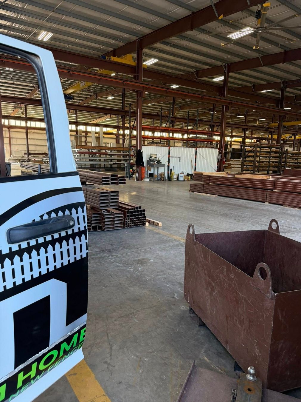 Warehouse interior with stacked metal beams, a blue vehicle edge, and large rusted metal bins on a concrete floor