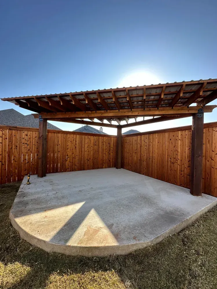 Covered wooden patio with concrete slab, fenced backyard, and bright sunlight casting shadows.