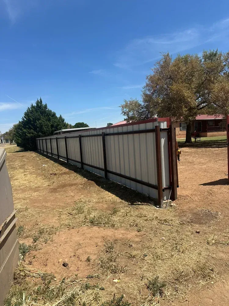 Long metal fence panels lined along a dirt lot under a blue sky, with trees and a building nearby