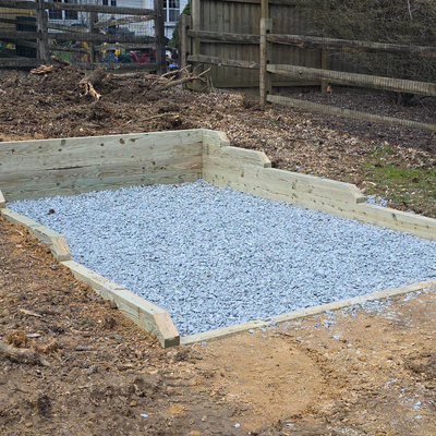 Gravel-filled square wooden border in a yard beside a black fence and stone wall.