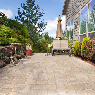 Sunny backyard patio with stone pavers, outdoor chairs, umbrella, and lush greenery beside a house