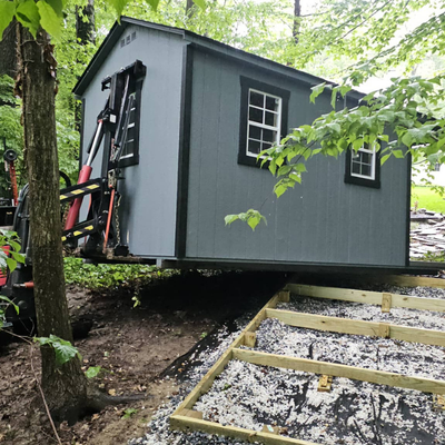 Small gray cabin or shed being moved through a wooded area, with a ladder and debris in front.