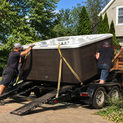 Two workers pushing a strapped hot tub onto a trailer in a sunny driveway.