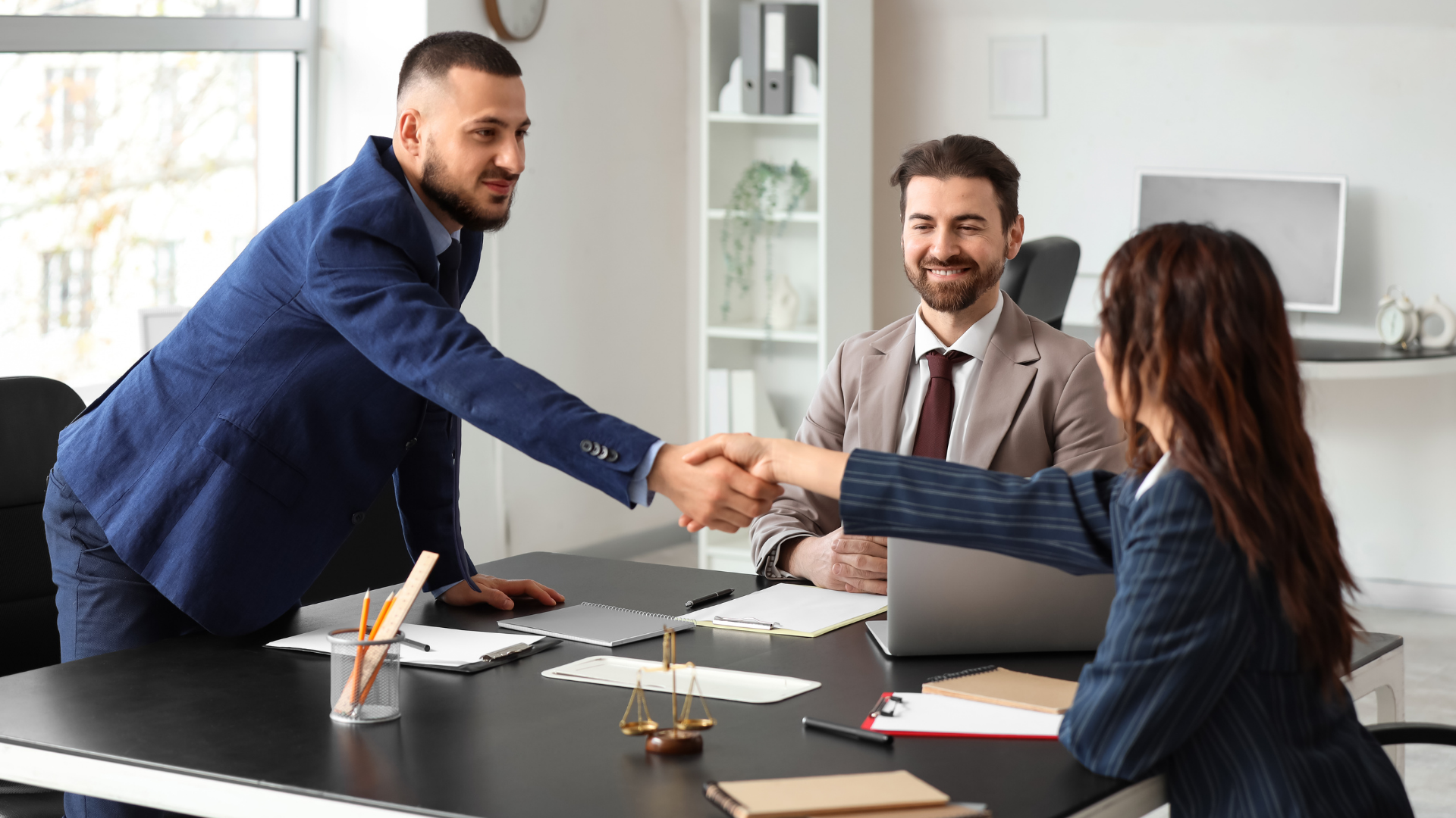 A professional in a blue blazer leans across a table to shake hands with a client while a colleague watches in an office.