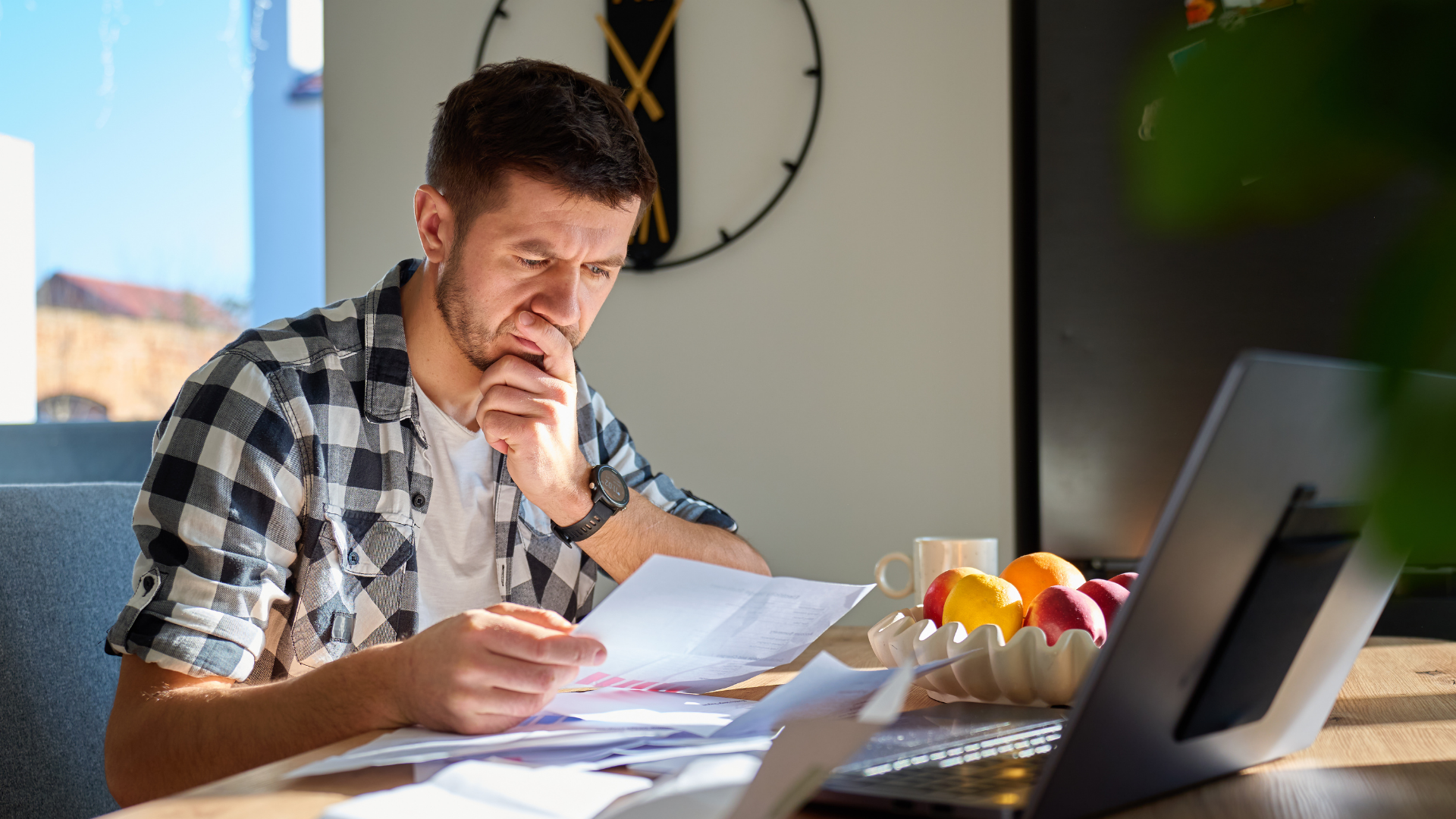 A person in a plaid shirt sits at a desk, looking concerned while reviewing paperwork near a laptop and a bowl of fruit.