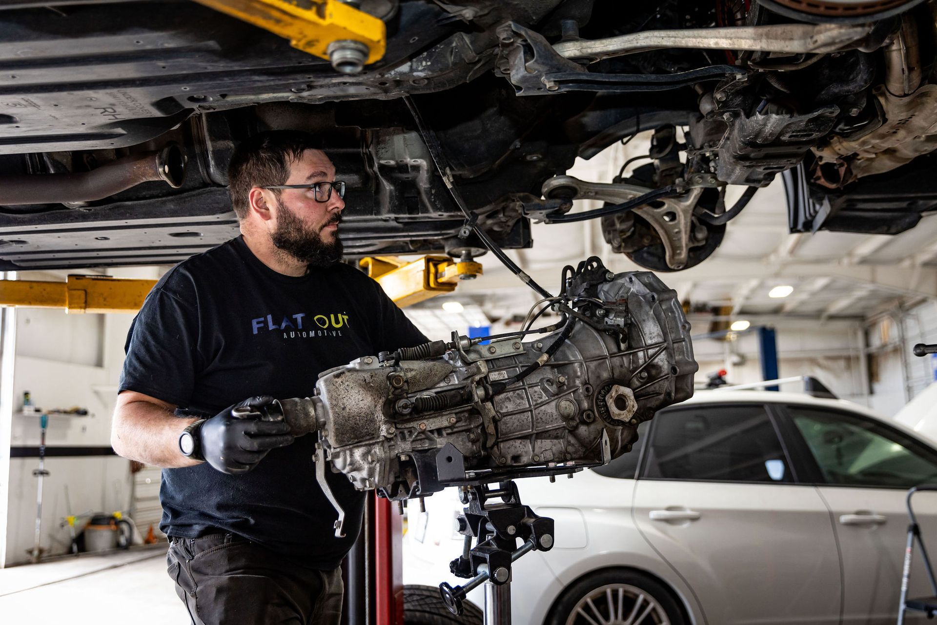 Mechanic removing car transmission, holding it with a lift in a garage. | Flatout Automotive