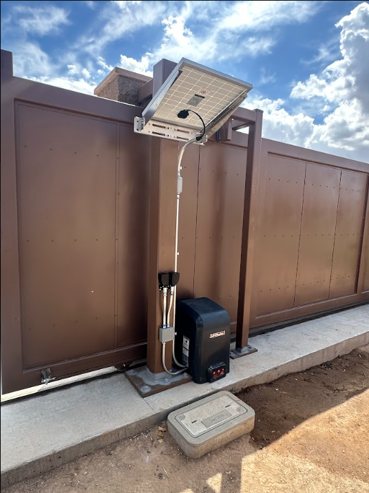 Outdoor lamp post with solar panel, battery box, and control unit beside a brown fence under a cloudy sky