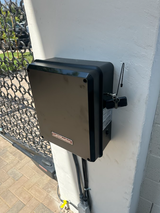 Black wall-mounted security/control box with antenna on a white outdoor wall beside a chain-link gate.