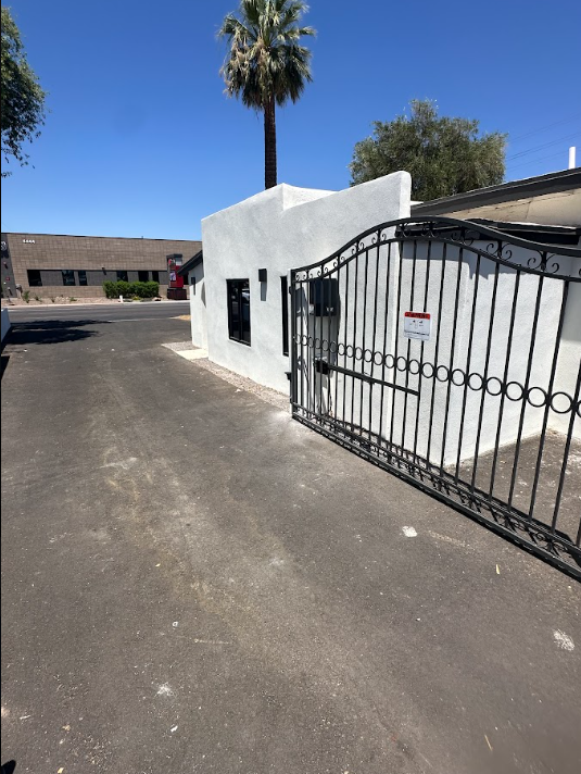 White stucco entrance with black wrought-iron gate beside a paved driveway under a palm tree.