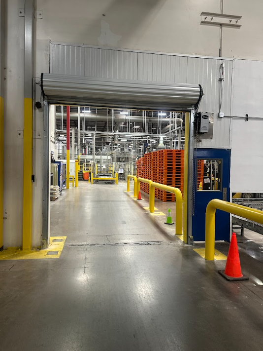 Industrial warehouse aisle with yellow railings, orange cones, and a partially open roll-up door