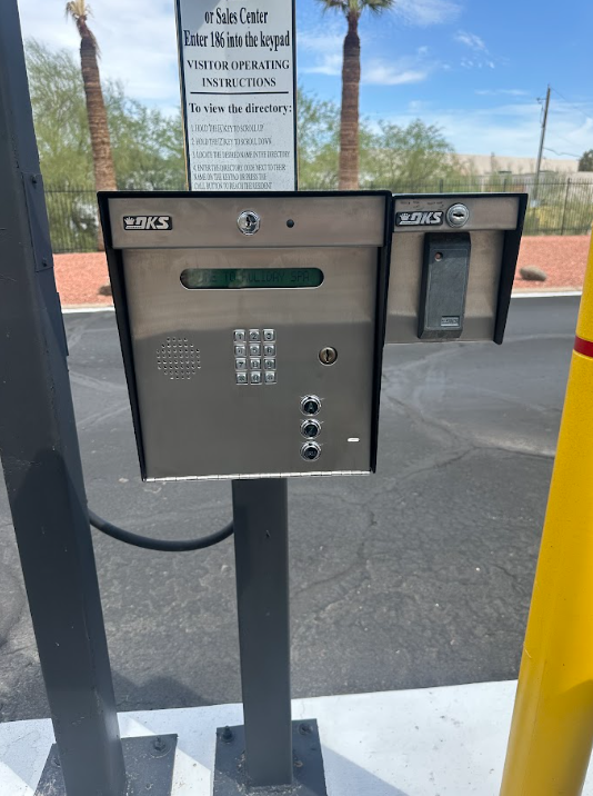 Outdoor security intercom and keypad mounted on a post, with a gate nearby under a bright sky.