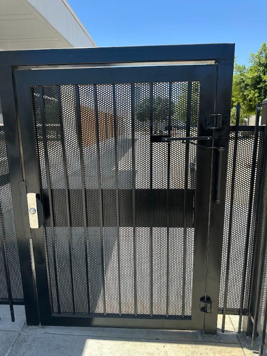 Black metal security gate with mesh panels and a latch, set in a fenced outdoor entrance.