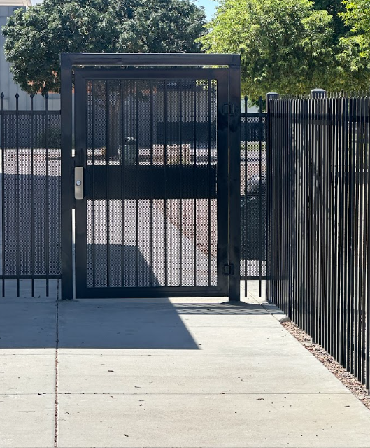 Black metal gate and fence beside a sunlit concrete walkway.