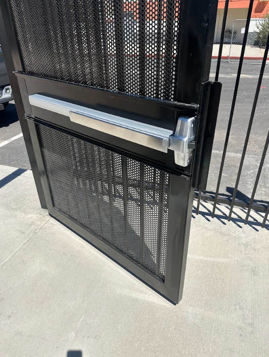 Black metal gate with a silver push bar on a sidewalk, partially open near a fenced entrance