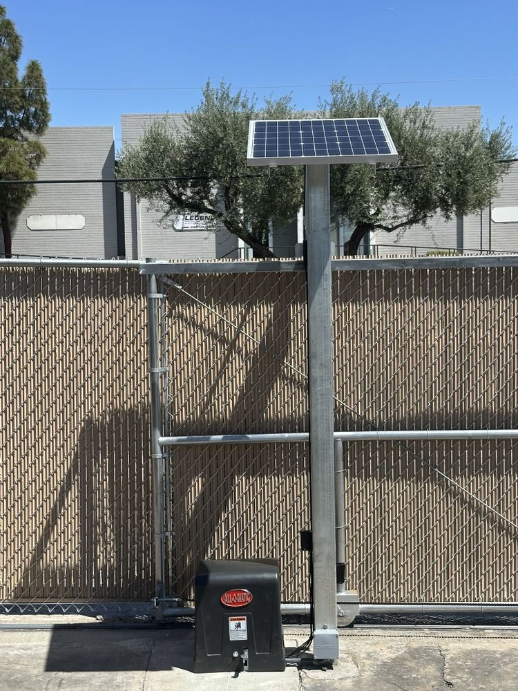 Solar-powered gate opener beside a wooden fence with a mounted panel and control box.