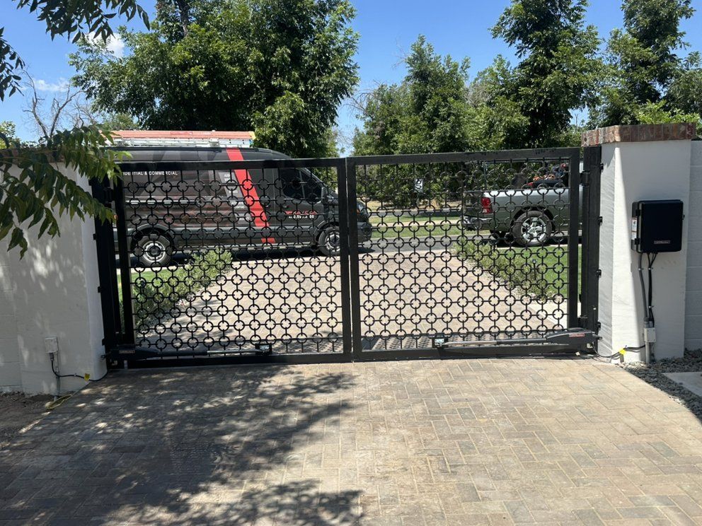 Black metal driveway gate at a sunny residential entrance, with cars and trees visible beyond.