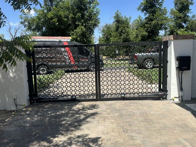 Black metal sliding gate at a driveway, with cars and trees visible beyond.