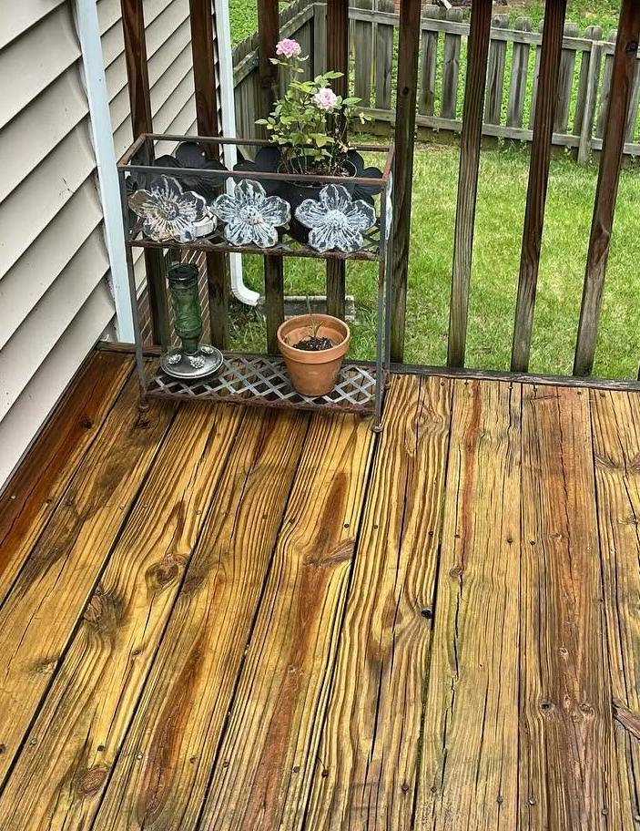 Wooden deck with a small plant stand, potted plant, and railing overlooking a grassy yard.