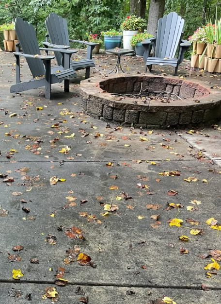 Backyard patio with Adirondack chairs around a stone fire pit, scattered fallen leaves on the ground
