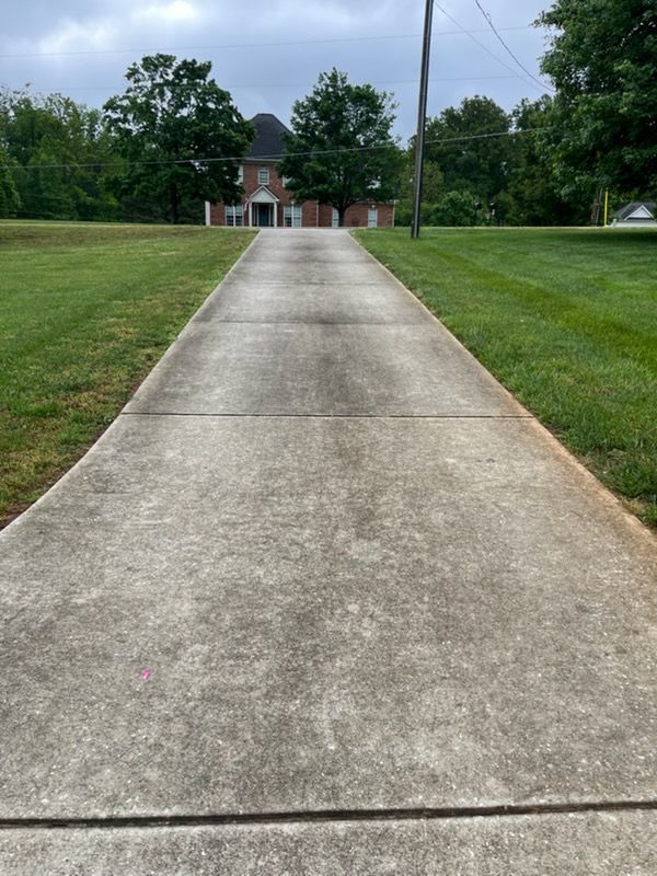 Long concrete driveway leading to a brick house with trees and lawn on a cloudy day