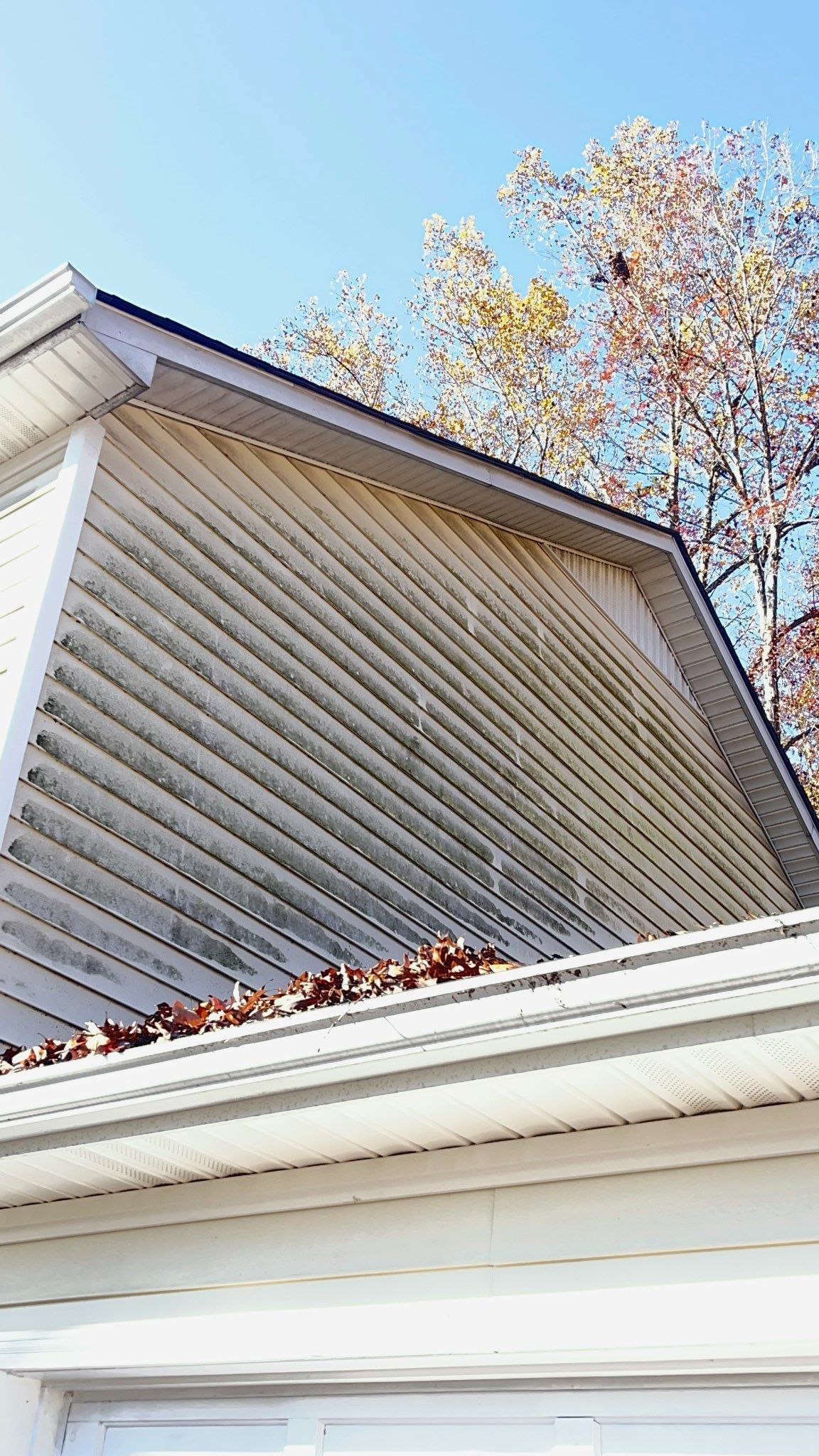 Close-up of a house gable with white siding and trim against a blue sky and autumn trees