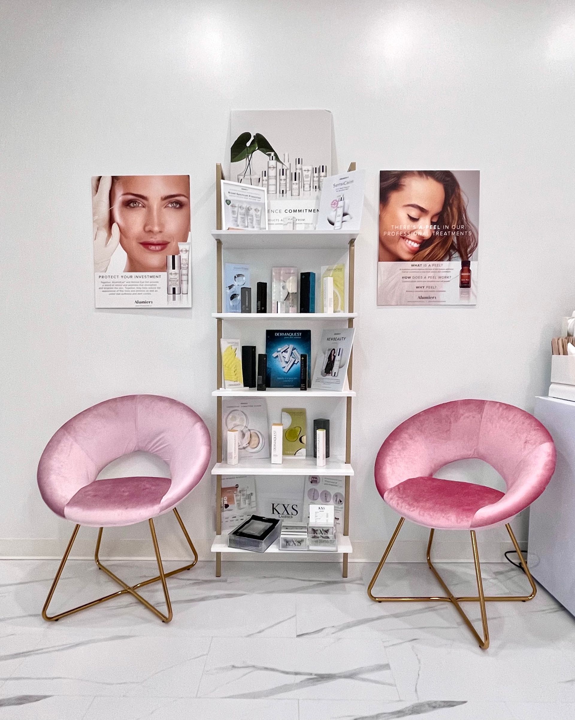 White salon waiting area with two pink chairs and a small shelf of beauty products and posters.