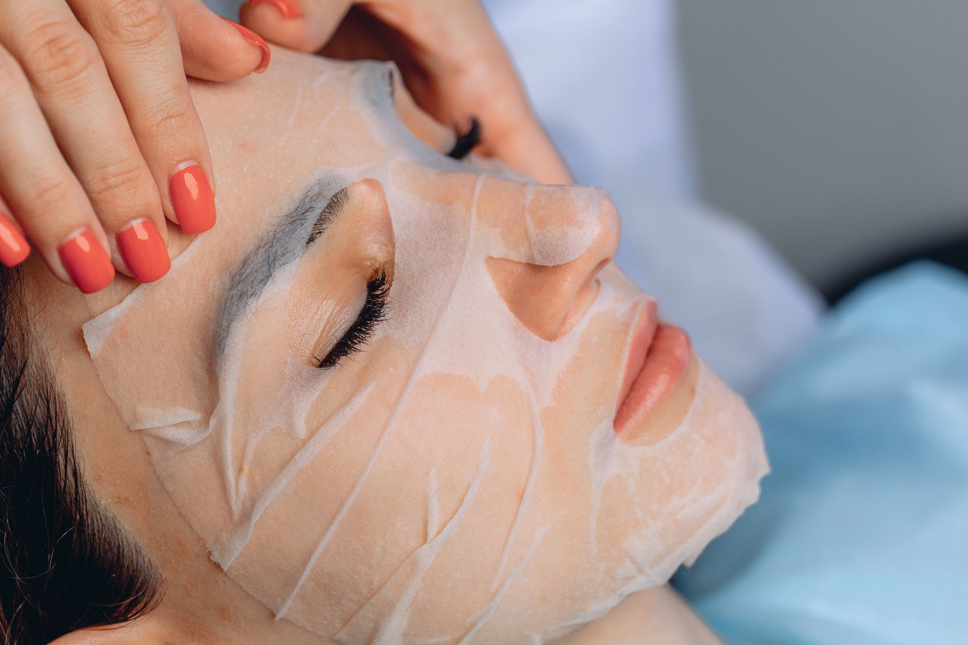 Woman receiving a facial treatment with a sheet mask and a hand massaging her forehead