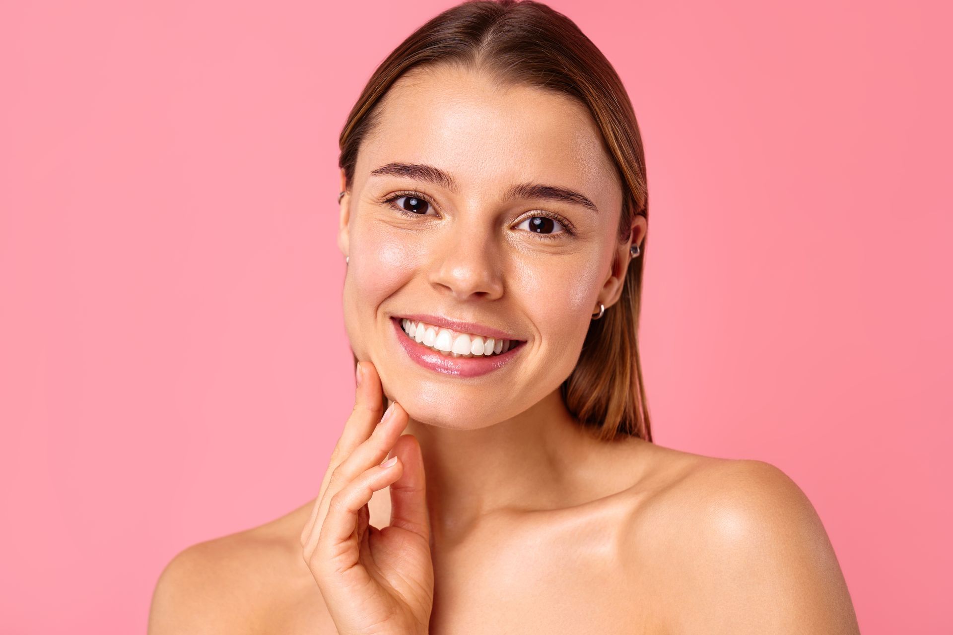 A woman is smiling and touching her face on a pink background.