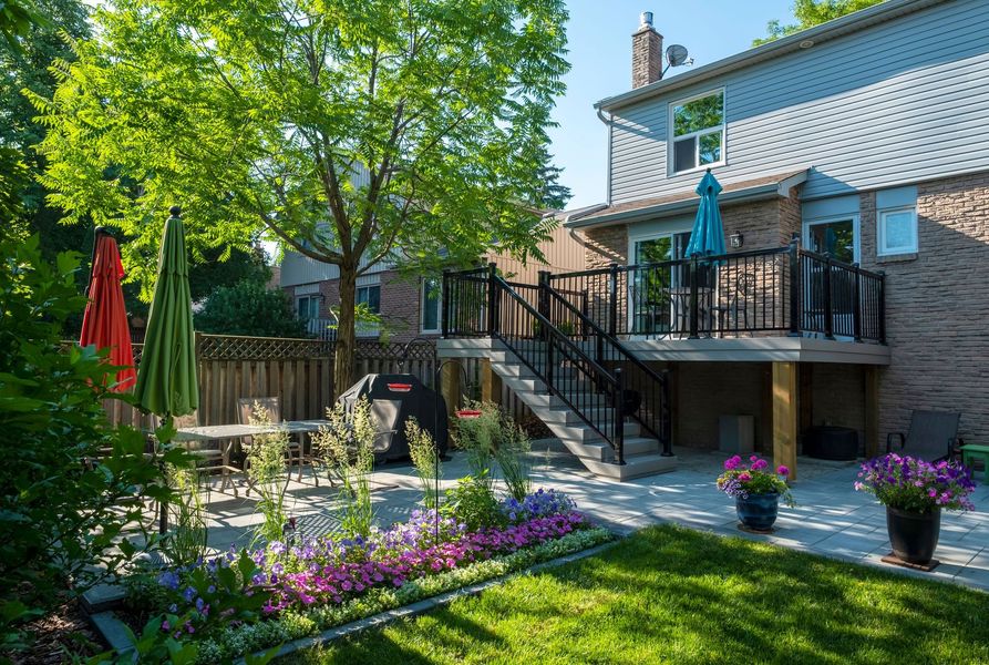 Backyard with deck, patio, and garden, featuring a house with brick siding, trees, and colorful umbrellas.