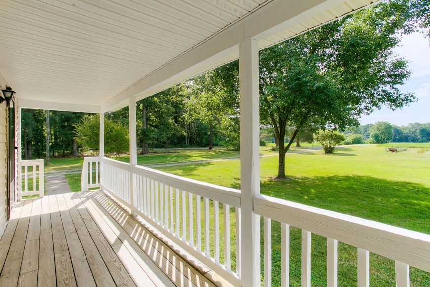 White porch with railing overlooking a sunny, grassy field and trees.
