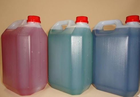 Tres jarras de plástico que contienen líquidos de color verde, lila y azul están alineadas en un estante.