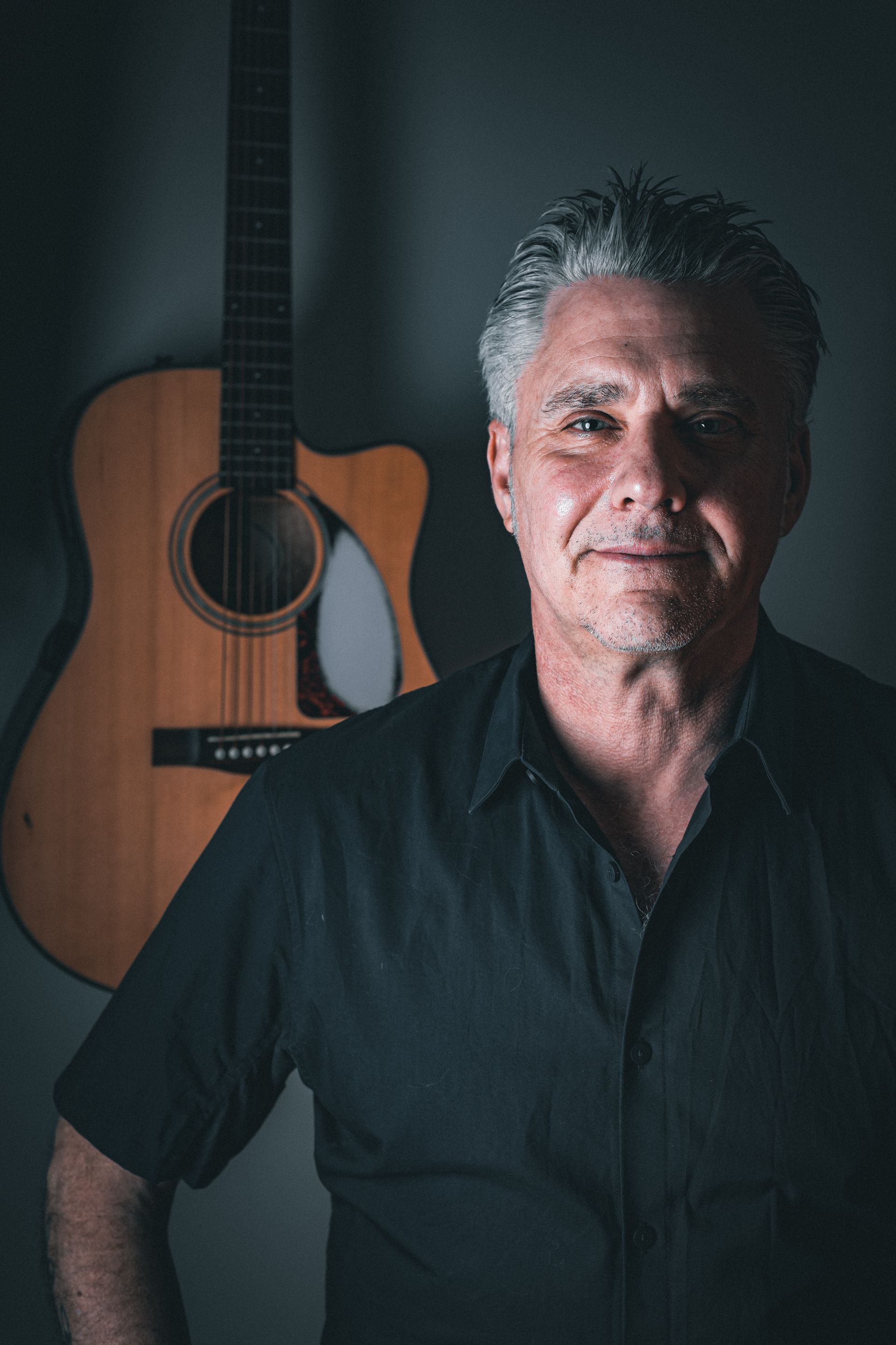 A man in a black shirt is standing in front of an acoustic guitar.