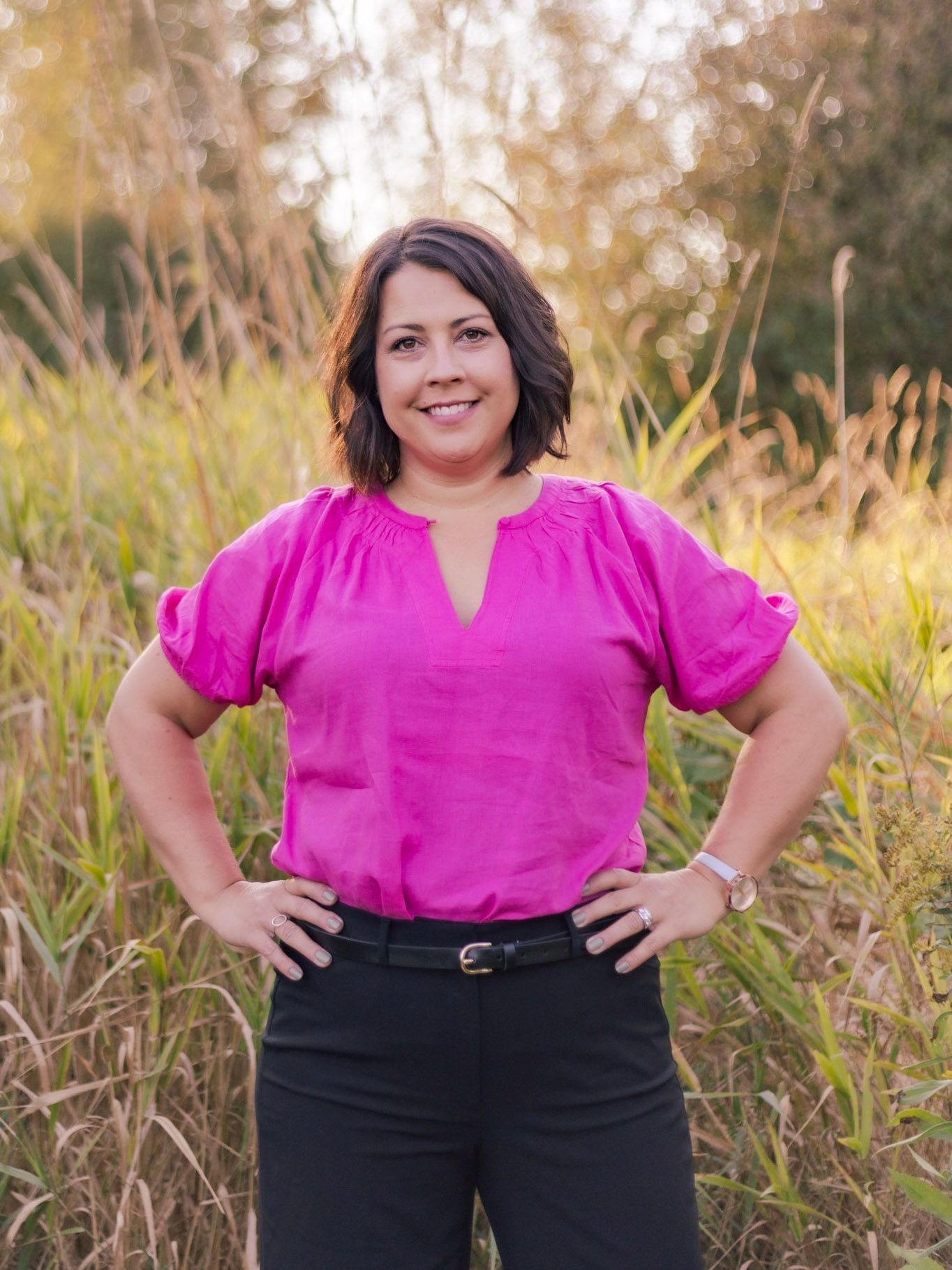 A woman in a pink shirt and black pants is standing in a field with her hands on her hips.