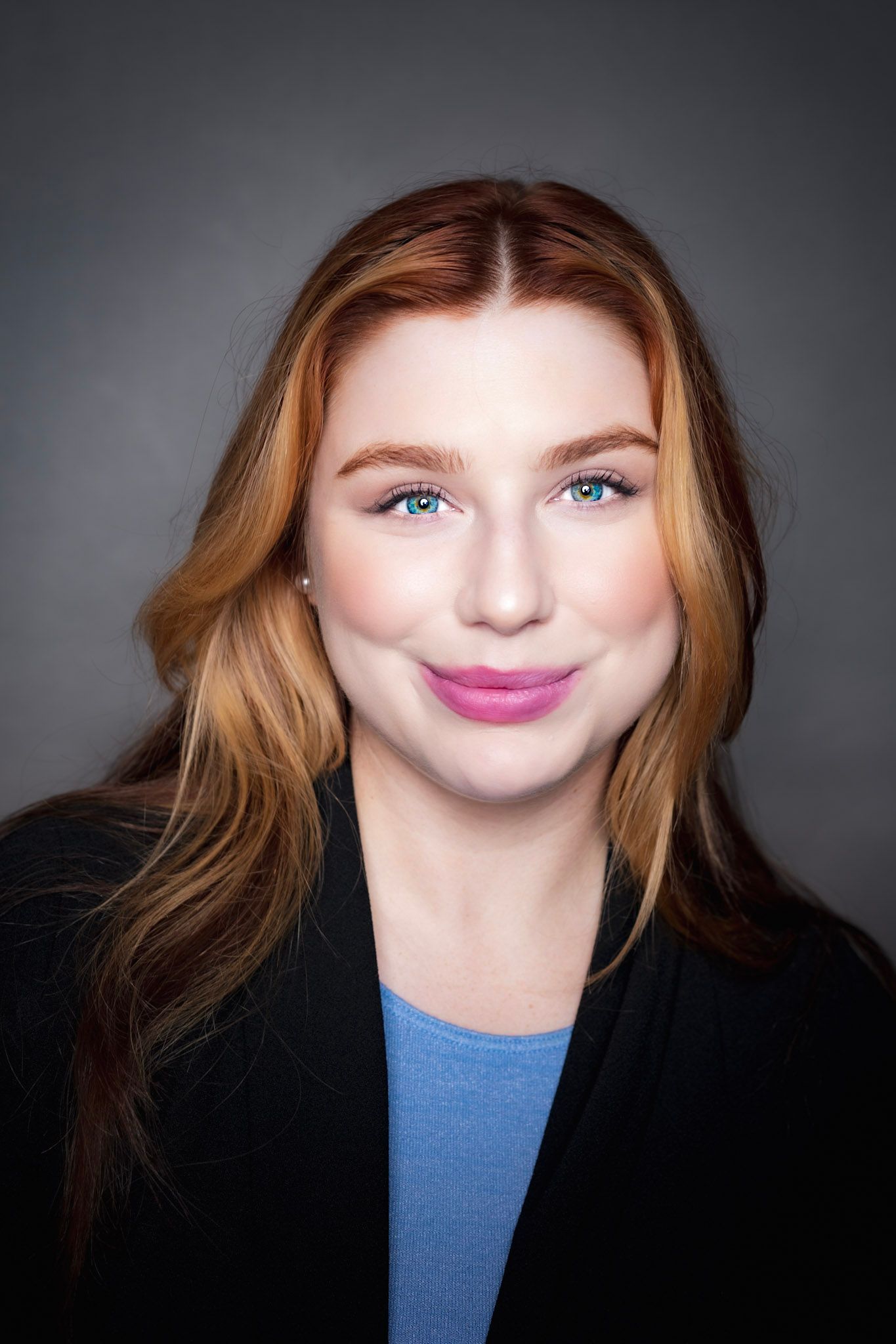 A woman with red hair and blue eyes is wearing a black jacket and a blue shirt.