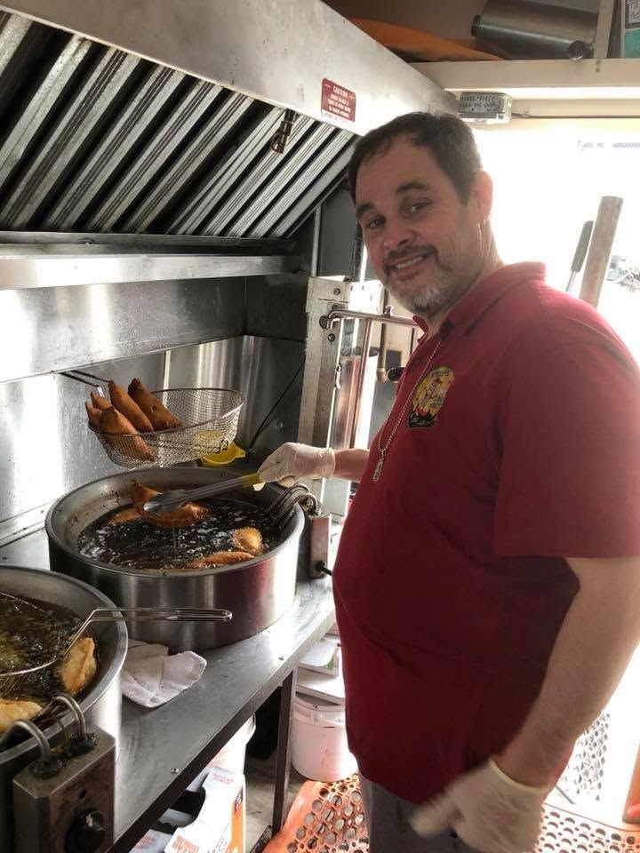 Man smiling beside steaming pots in a commercial kitchen, tending food on a stovetop.