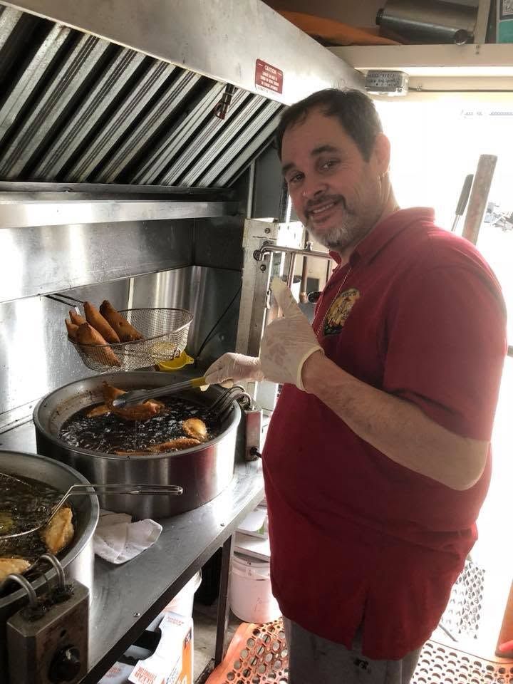Man in a red shirt and gloves cooking at a busy kitchen stove, holding a skewer and smiling.