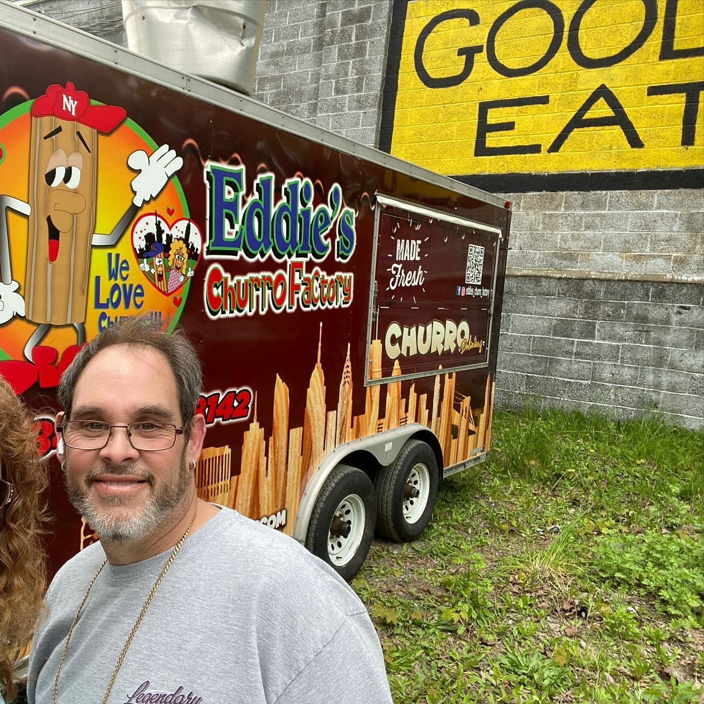 Man smiling in front of an Eddie’s Churro trailer with a large yellow “GOOD EATS” sign behind it