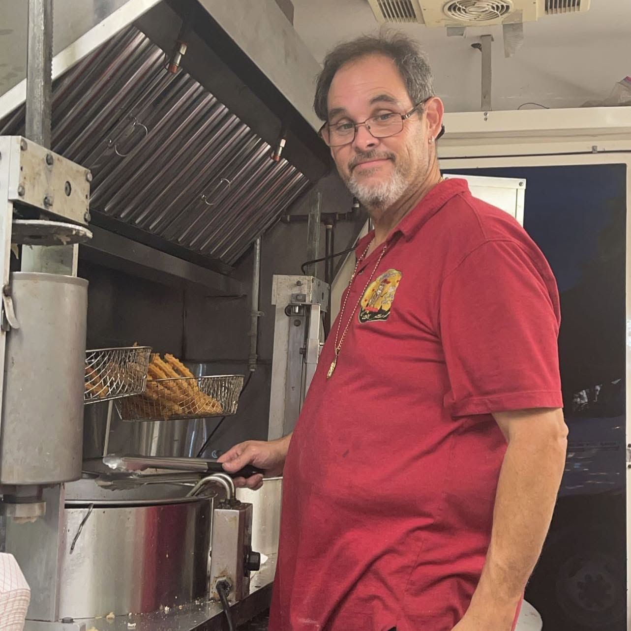 Man in red shirt standing by a grill in a kitchen, holding a pot and smiling.