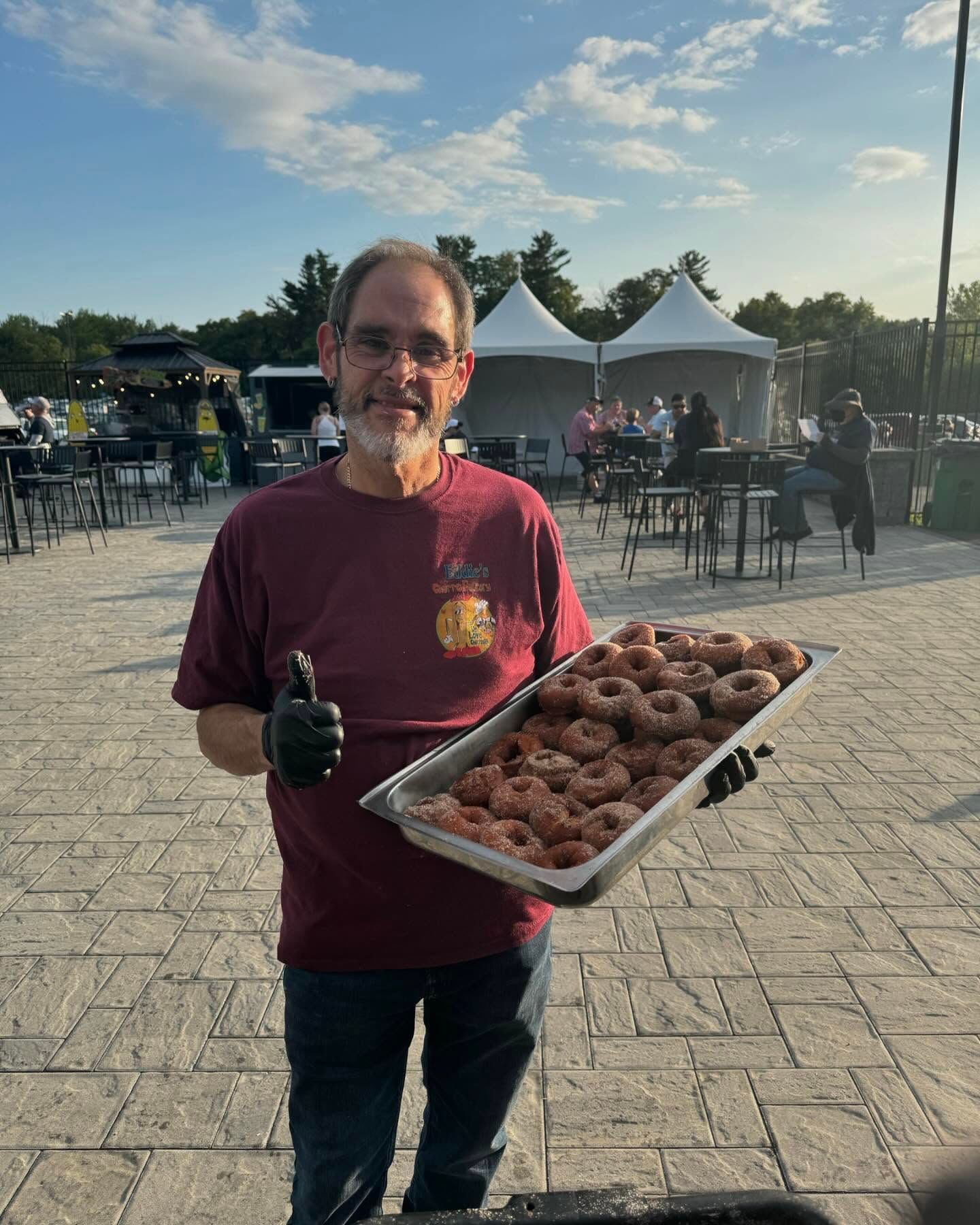 Man holding a tray of donuts outdoors at an event, with tents and tables in the background.