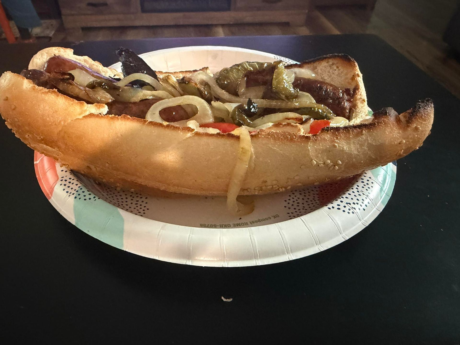 Long sandwich with grilled meat and onions on a white paper plate on a dark table