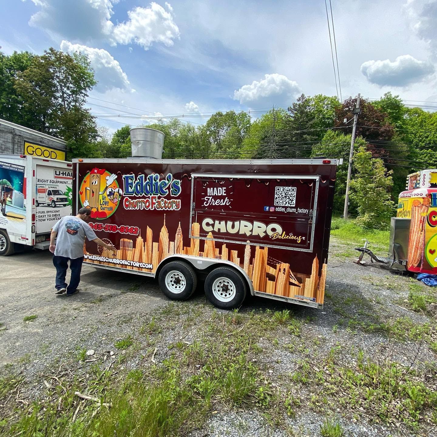 Food truck trailer with a brown churros-themed wrap parked on gravel, with a person standing beside it.
