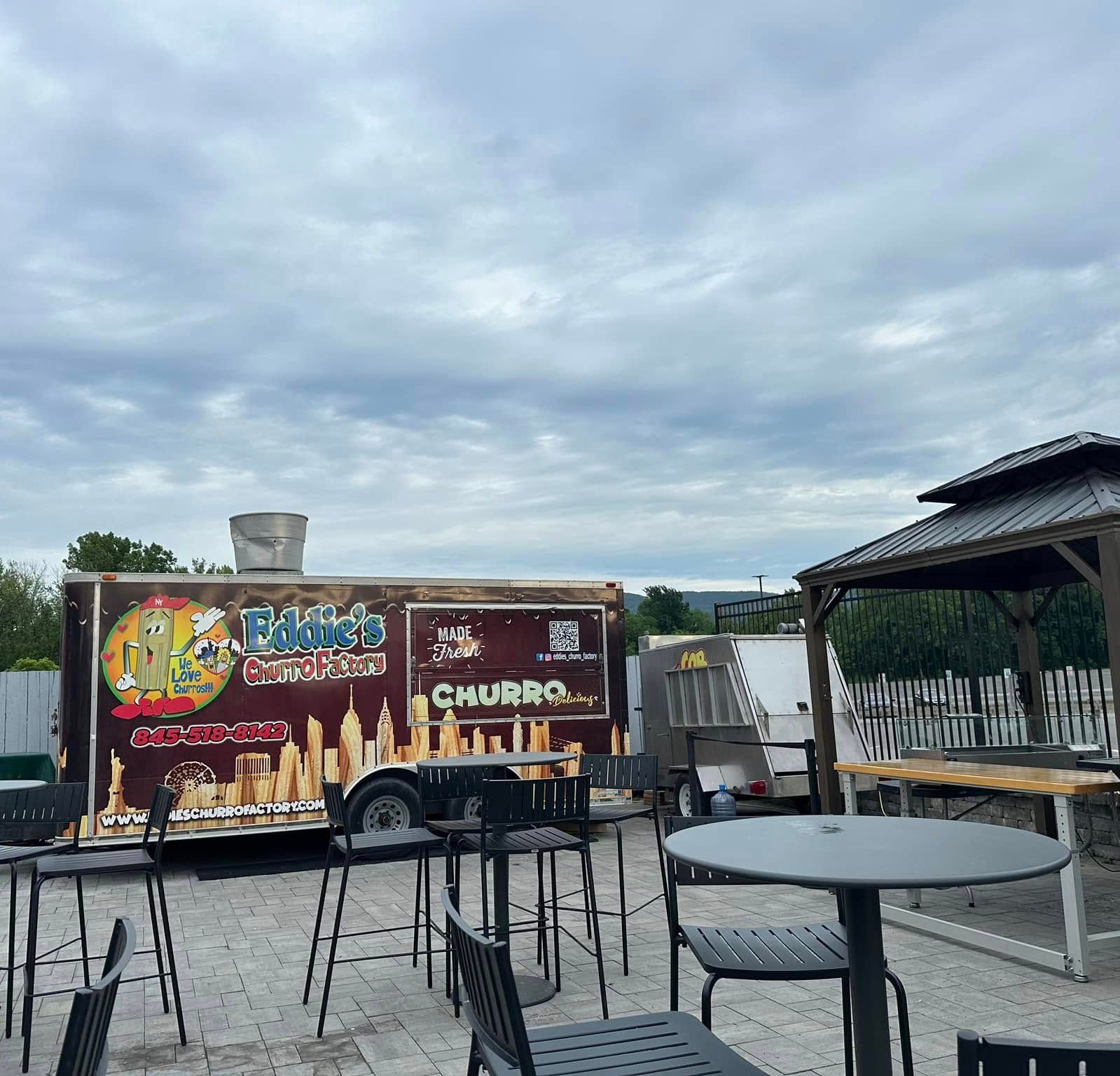 Food truck with colorful menu signs parked beside an outdoor patio under a cloudy sky
