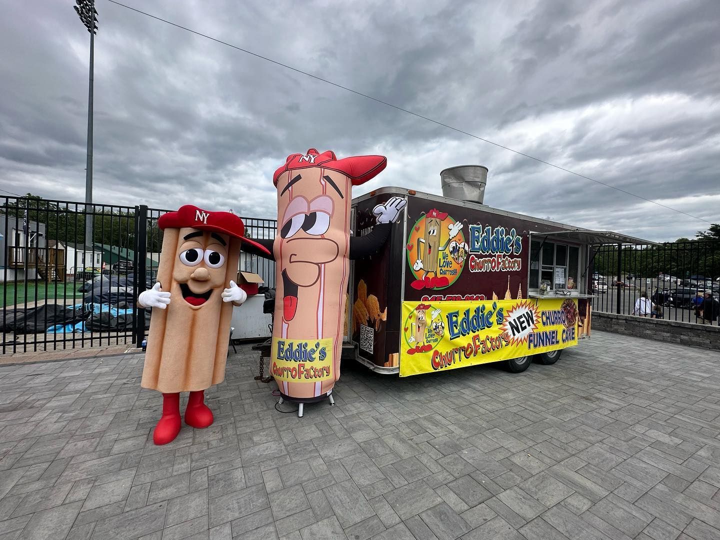 Two cartoon food mascots beside a colorful food truck in a paved parking lot under cloudy skies