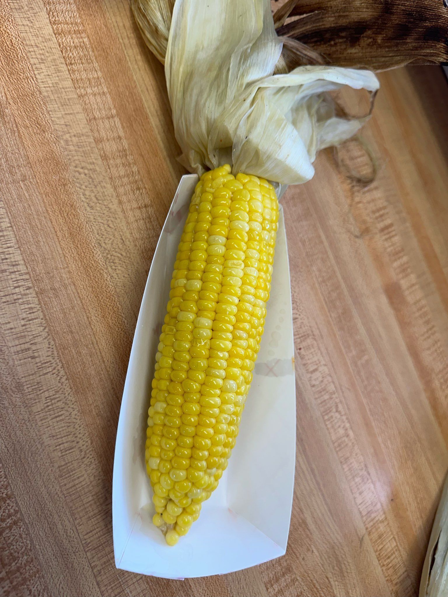 A yellow ear of corn in a white wrapper on a wooden table, with husk and silk at the top