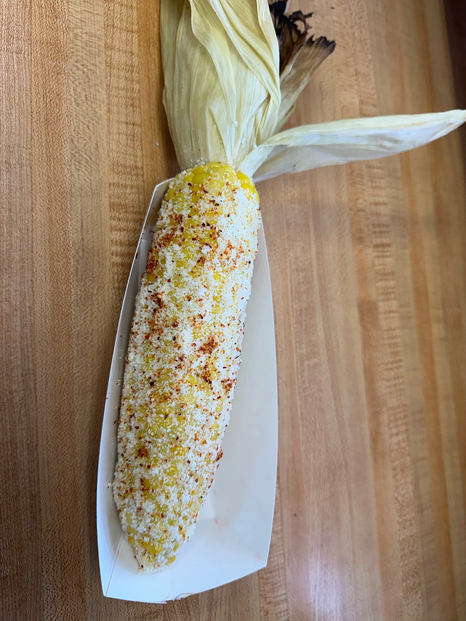 Corn cob on a wooden board, partly husked and seasoned with red and yellow spices.