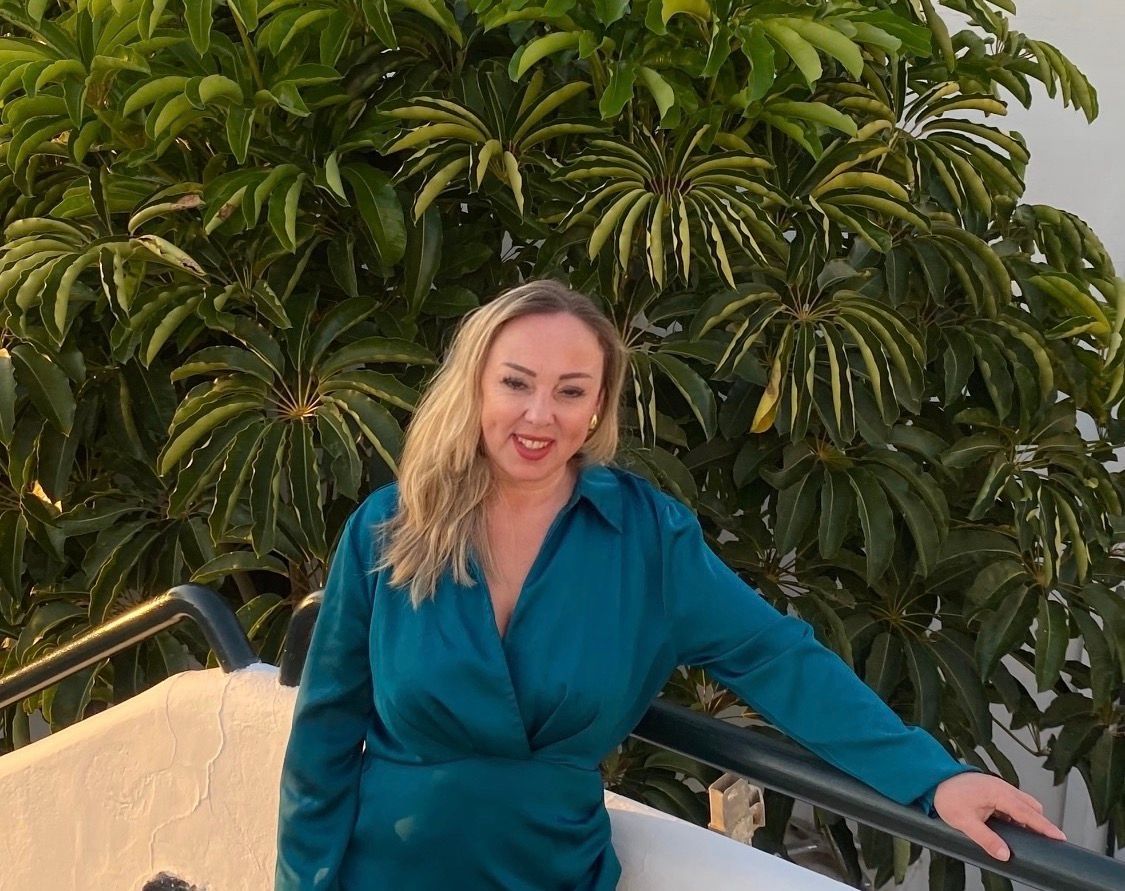 Vrouw in een turquoise blouse lacht op een balkon met weelderig groen op de achtergrond.